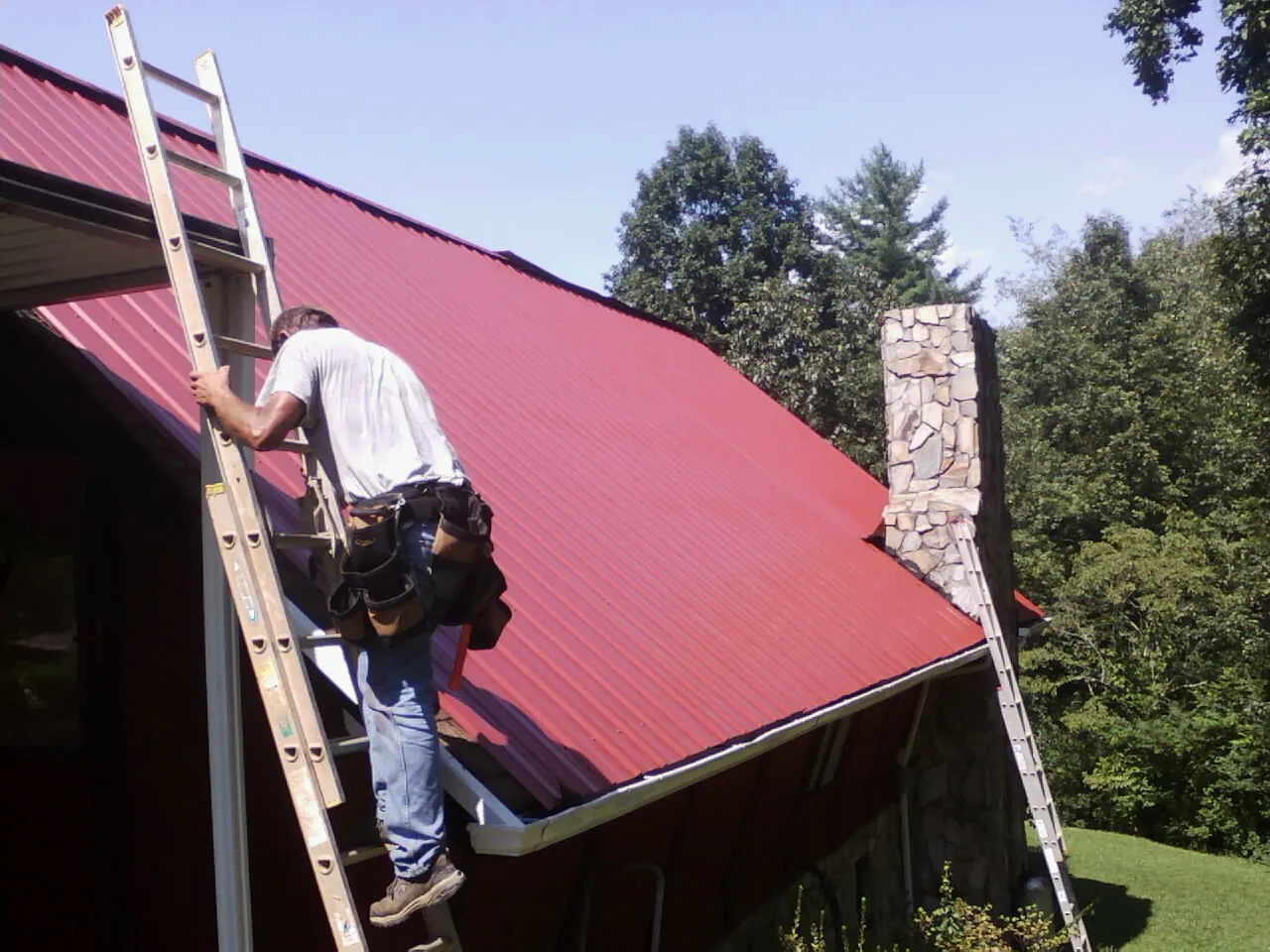 Roofer on a ladder beside a red metal roof and stone chimney — Soffit & Fascia Repair by Roofing Brothers in Gambell